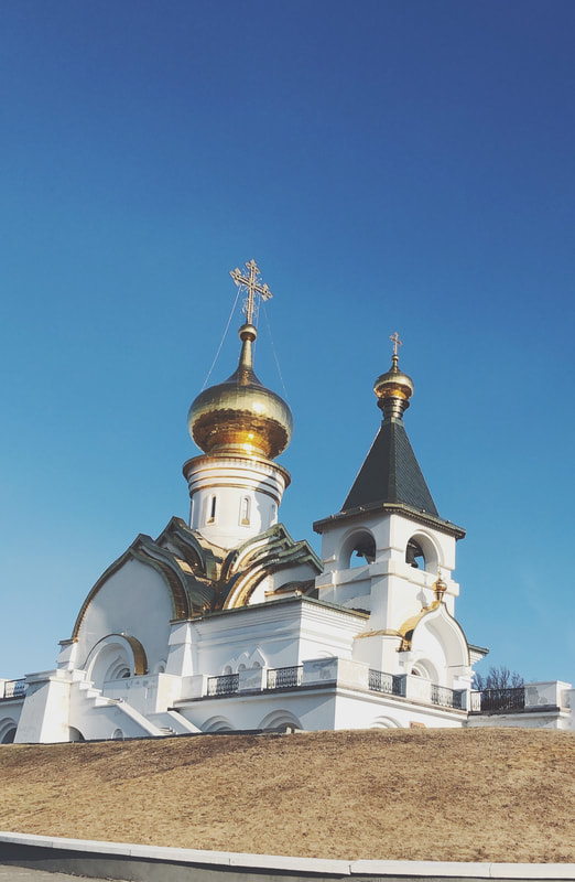 An image of an Eastern Orthodox Church in Russia is shown. The church is white and gold. The sky behind it is blue and the field in front is dry.