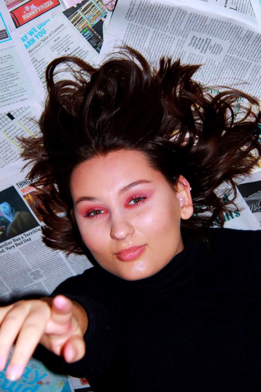 A woman points at the camera while laying down on a pile of magazines. She is wearing a black turtleneck and pink makeup. The image is a flash photo.