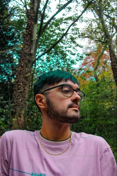 A camera angled from below shows a man looking down at the ground through his glasses. His hair appears to be turquoise and the forest behind him is very vibrant.