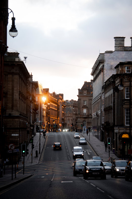 An image of sunset is shown in the city center of Newcastle Upon Tyne, United Kingdom. Cars are shown driving in both directions and old buildings surround the street. The photo has a dark tone to it.