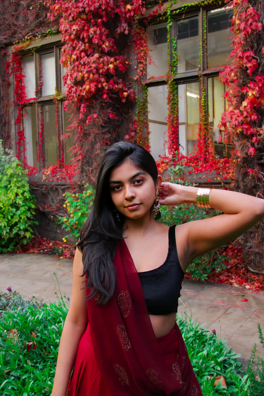A woman is wearing a red sari in front of an ivy covered wall. The wall is mostly red whereas the surrounding plants are green.